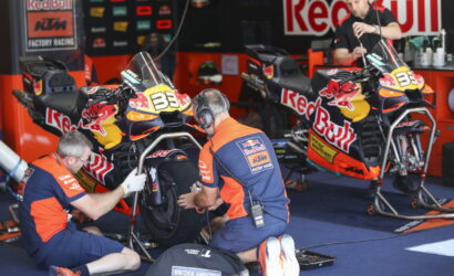 Two Red Bull KTM factory racing motorcycles are on stands in a busy pit garage as crew members service the bikes and check tires behind them.