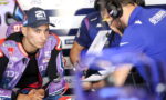 Moto racer in purple Pramac gear sits in the pit, talking with a crew member wearing a headset and clipboard nearby