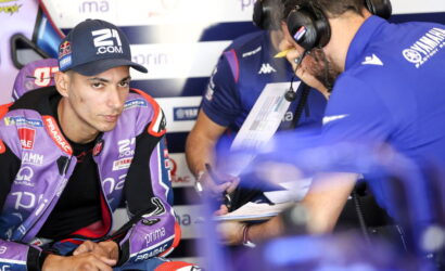 Moto racer in purple Pramac gear sits in the pit, talking with a crew member wearing a headset and clipboard nearby