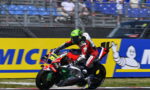 Motorcycle racer in red‑white gear leans into a sharp turn on a track, helmet neon green and number 36 visible on the bike, sponsors on the suit and bike nearby.