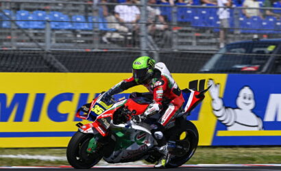 Motorcycle racer in red‑white gear leans into a sharp turn on a track, helmet neon green and number 36 visible on the bike, sponsors on the suit and bike nearby.