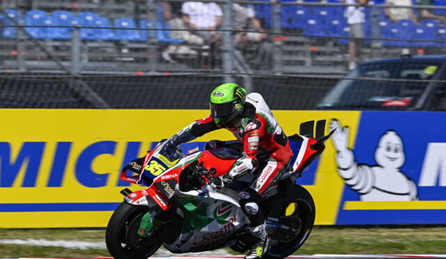 Motorcycle racer in red‑white gear leans into a sharp turn on a track, helmet neon green and number 36 visible on the bike, sponsors on the suit and bike nearby.