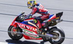Racing motorcycle rider leaning into a turn on a red Honda race bike with sponsor logos on a track.