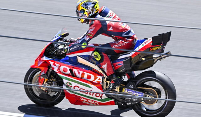 Racing motorcycle rider leaning into a turn on a red Honda race bike with sponsor logos on a track.