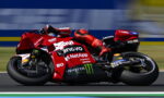 Red racing motorcycle in a sharp left turn with riders in red gear; sponsors like Lenovo, NetApp, Monster Energy visible on the bike's bodywork, motion blur in background.