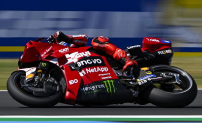 Red racing motorcycle in a sharp left turn with riders in red gear; sponsors like Lenovo, NetApp, Monster Energy visible on the bike's bodywork, motion blur in background.