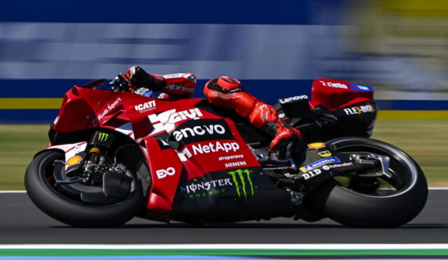 Red racing motorcycle in a sharp left turn with riders in red gear; sponsors like Lenovo, NetApp, Monster Energy visible on the bike's bodywork, motion blur in background.