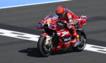 Motorcycle racer in red suit leaning into a turn on a red Ducati racing bike with sponsor logos visible on track surface behind him.