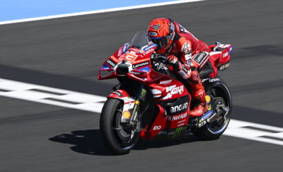 Motorcycle racer in red suit leaning into a turn on a red Ducati racing bike with sponsor logos visible on track surface behind him.