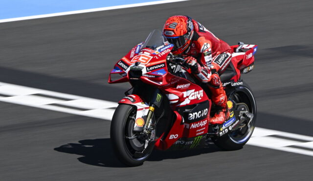 Motorcycle racer in red suit leaning into a turn on a red Ducati racing bike with sponsor logos visible on track surface behind him.