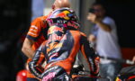 Moto racer in an orange KTM suit sits on a bike, rear view, helmet branded with Red Bull graphics and sprint-ready stance.