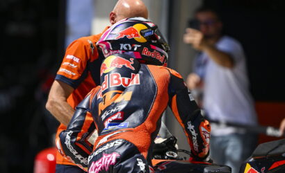 Moto racer in an orange KTM suit sits on a bike, rear view, helmet branded with Red Bull graphics and sprint-ready stance.