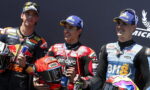 Three motorcycle racers stand on a podium, smiling, each holding a helmet and a drink in their hands.