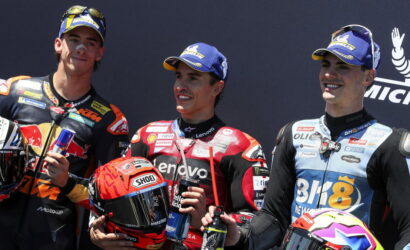Three motorcycle racers stand on a podium, smiling, each holding a helmet and a drink in their hands.