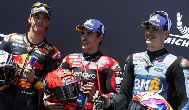 Three motorcycle racers stand on a podium, smiling, each holding a helmet and a drink in their hands.