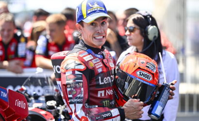 Moto racer in red racing suit holds orange Shoei helmet, smiling at camera with pit crew in background. Marked with Michelin cap and sponsor logos.