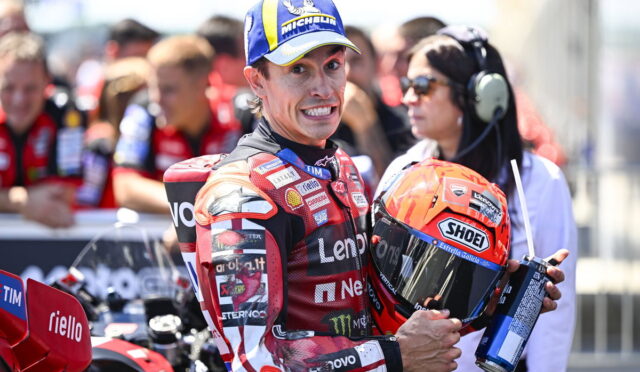 Moto racer in red racing suit holds orange Shoei helmet, smiling at camera with pit crew in background. Marked with Michelin cap and sponsor logos.