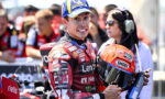 MotoGP rider in a red Lenovo racing suit holding an orange Shoei helmet, smiles at the camera with pit crew in the blurred background.