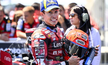 MotoGP rider in a red Lenovo racing suit holding an orange Shoei helmet, smiles at the camera with pit crew in the blurred background.