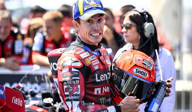 MotoGP rider in a red Lenovo racing suit holding an orange Shoei helmet, smiles at the camera with pit crew in the blurred background.