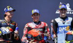 Three motorcycle racers on a podium pose with drinks; center rider wears red racing suit and holds an orange helmet and a water bottle filled with blue liquid on his chest.