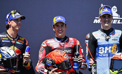 Three motorcycle racers on a podium pose with drinks; center rider wears red racing suit and holds an orange helmet and a water bottle filled with blue liquid on his chest.