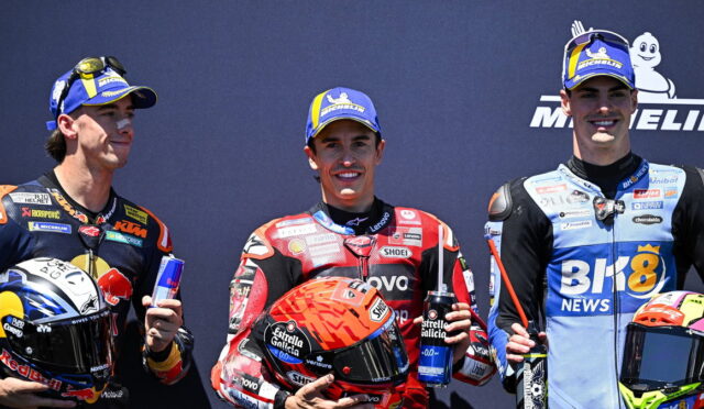 Three motorcycle racers on a podium pose with drinks; center rider wears red racing suit and holds an orange helmet and a water bottle filled with blue liquid on his chest.