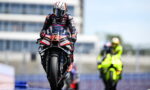 Moto racer on a sport bike racing toward the camera, helmeted rider in red-blue suit on a track with a neon-yellow rider blurred in the background.