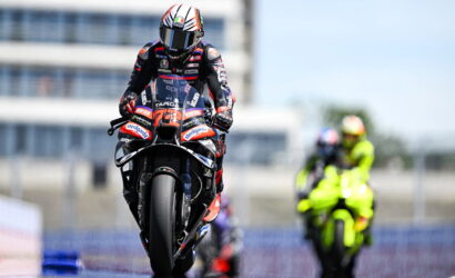 Moto racer on a sport bike racing toward the camera, helmeted rider in red-blue suit on a track with a neon-yellow rider blurred in the background.
