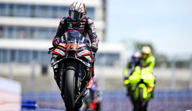 Moto racer on a sport bike racing toward the camera, helmeted rider in red-blue suit on a track with a neon-yellow rider blurred in the background.
