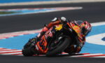 Motorcycle racer in an orange suit leaning into a sharp turn on a race track.