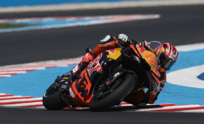 Motorcycle racer in an orange suit leaning into a sharp turn on a race track.