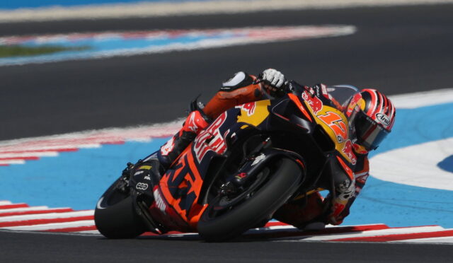 Motorcycle racer in an orange suit leaning into a sharp turn on a race track.