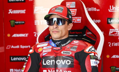 Rider in a red Lenovo racing suit sits in a branded pit area wearing a cap and mirrored sunglasses.