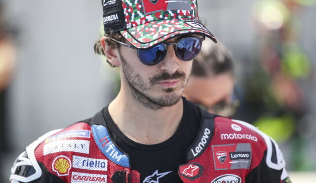 Male motorcyclist with a red camouflage cap and mirrored sunglasses, wearing a red racing suit with sponsor logos.
