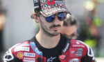 Male motorcyclist wearing a camouflage cap and blue mirrored sunglasses, red sponsor-laden racing suit.