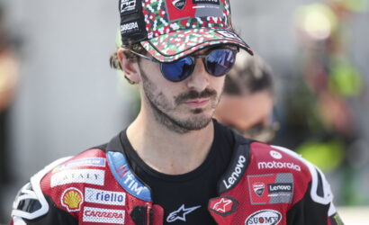 Male motorcyclist wearing a camouflage cap and blue mirrored sunglasses, red sponsor-laden racing suit.
