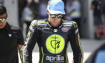 MotoGP rider in a black racing suit with neon trim, wearing a blue Trackhouse cap and sunglasses in a pit area