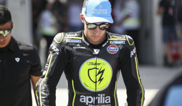 MotoGP rider in a black racing suit with neon trim, wearing a blue Trackhouse cap and sunglasses in a pit area