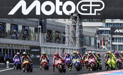 Motorcycle racers line up at the starting grid under a large MotoGP banner at a race track