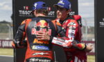 Two MotoGP riders in racing suits share a hug on the track, with sponsor logos visible (Red Bull, Michelin).