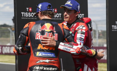 Two MotoGP riders in racing suits share a hug on the track, with sponsor logos visible (Red Bull, Michelin).