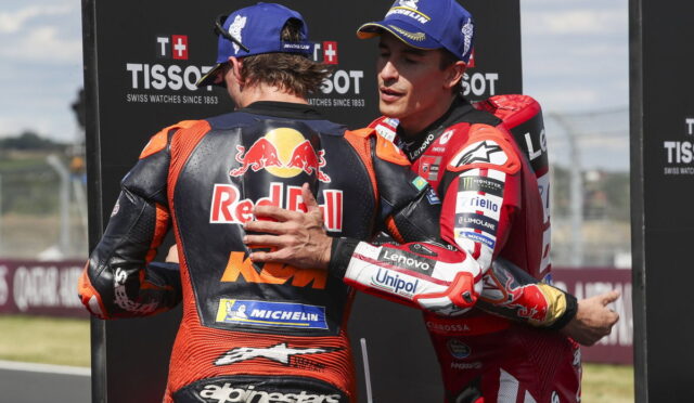 Two MotoGP riders in racing suits share a hug on the track, with sponsor logos visible (Red Bull, Michelin).