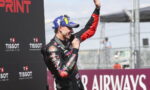 MotoGP-style racer in racing suit on podium, raising a trophy and hand in victory against sponsor backdrop