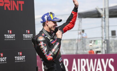 MotoGP-style racer in racing suit on podium, raising a trophy and hand in victory against sponsor backdrop