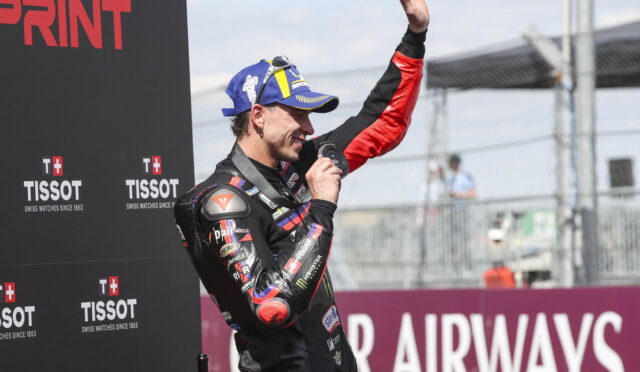 MotoGP-style racer in racing suit on podium, raising a trophy and hand in victory against sponsor backdrop
