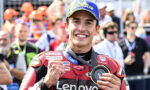 MotoGP rider in red Lenovo racing suit celebrates victory, flashing a fist and displaying a medal as a crowd watches in the background.