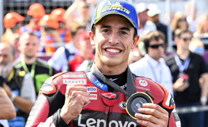 MotoGP rider in red Lenovo racing suit celebrates victory, flashing a fist and displaying a medal as a crowd watches in the background.