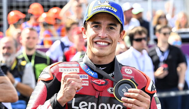 MotoGP rider in red Lenovo racing suit celebrates victory, flashing a fist and displaying a medal as a crowd watches in the background.