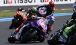 MotoGP riders lean into a corner on a race track, purple foreground bike #77 with sponsor decals.